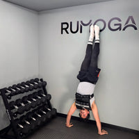 Person doing a handstand in a gym with 'RUMOGA' branding on the wall wearing a weight vest