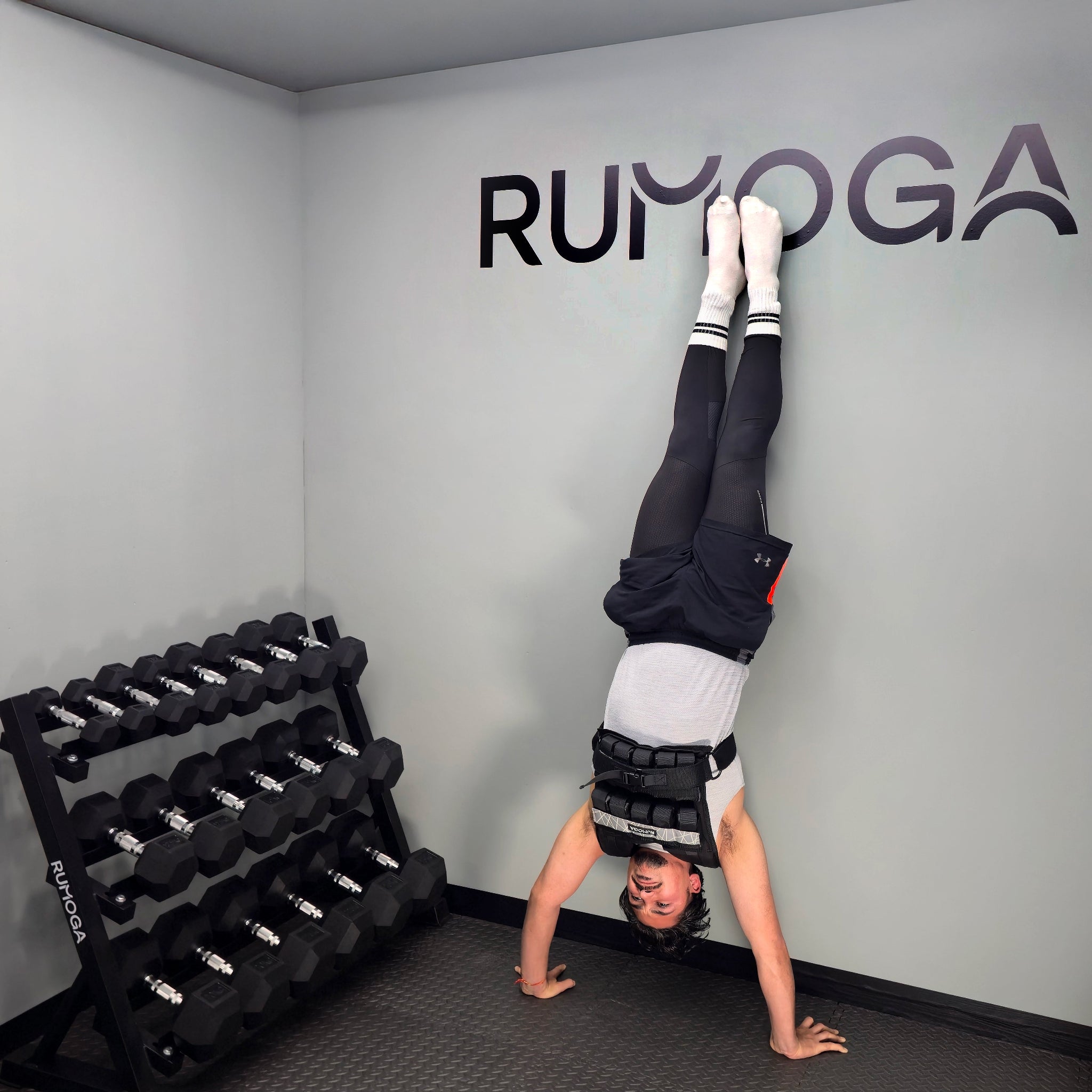 Person doing a handstand in a gym with 'RUMOGA' branding on the wall wearing a weight vest